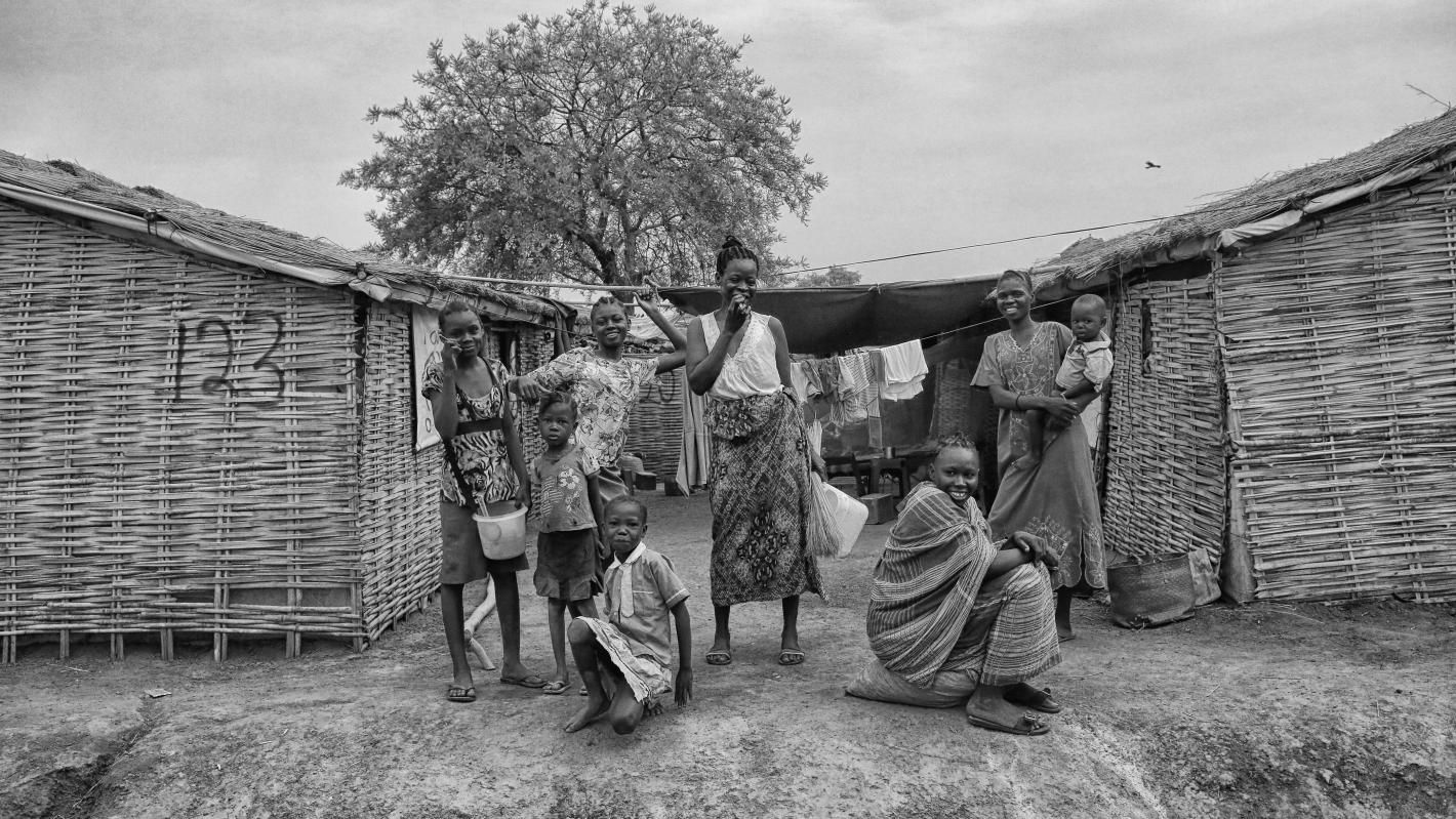 A group of adults and children stand and sit outside two thatched huts made of woven sticks, with laundry hanging on a line and a large tree in the background.