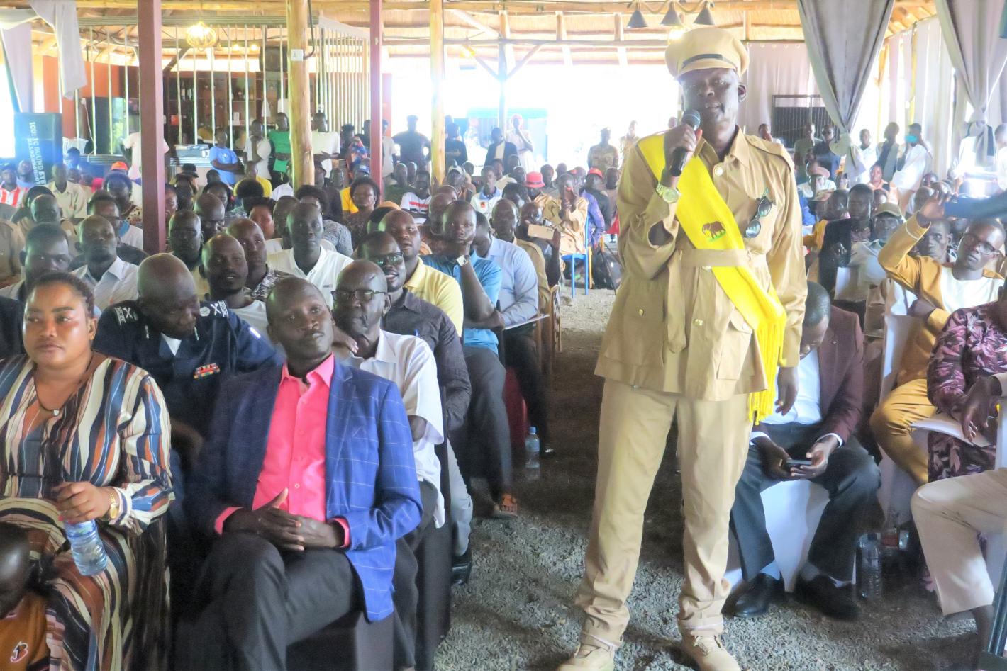 A military person standing in a yellow uniform wearing a yellow sash is speaking into a microphone to a large room of people seated in several rows behind them.