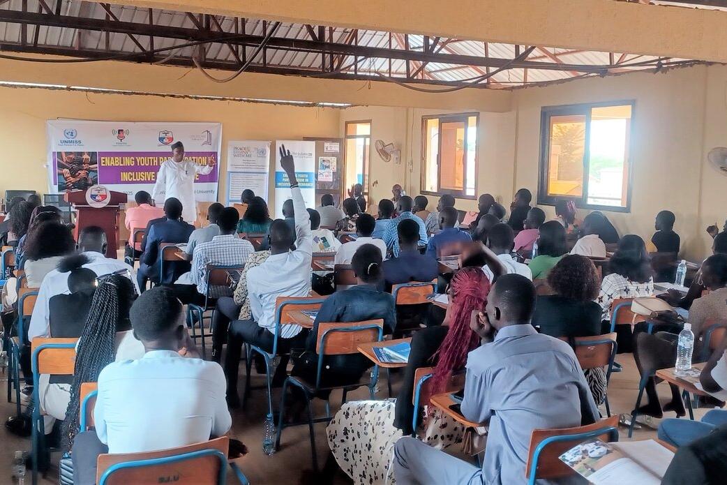 A classroom-style setting with many people seated at desks facing a speaker at the front, where banners and a presentation on youth empowerment are displayed.