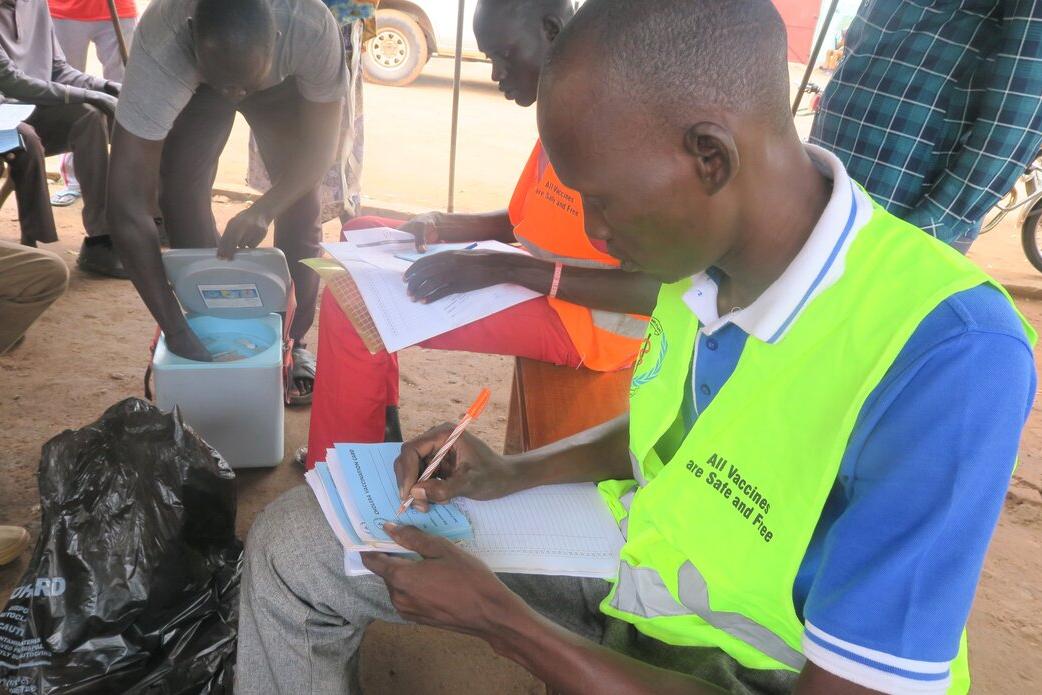 Health worker wearing a bright vest writes in a register during a vaccination campaign, with a cooler box and other people seated nearby.