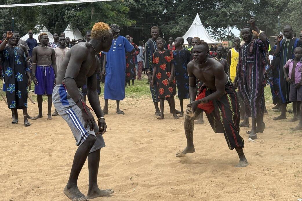 Two individuals engaged in a traditional wrestling match on sandy ground, surrounded by a crowd wearing colorful patterned clothing, with white tents and trees in the background.