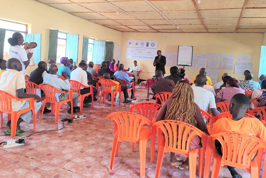 A community meeting in a room with orange plastic chairs arranged in rows, participants seated and listening to a speaker at the front near posters and a banner on the wall.